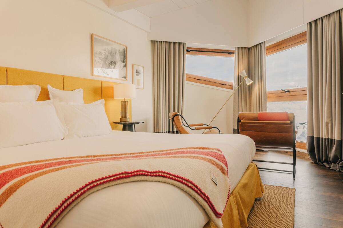 A hotel room with a bed, a chair, a desk, a lamp, and a window with curtains.