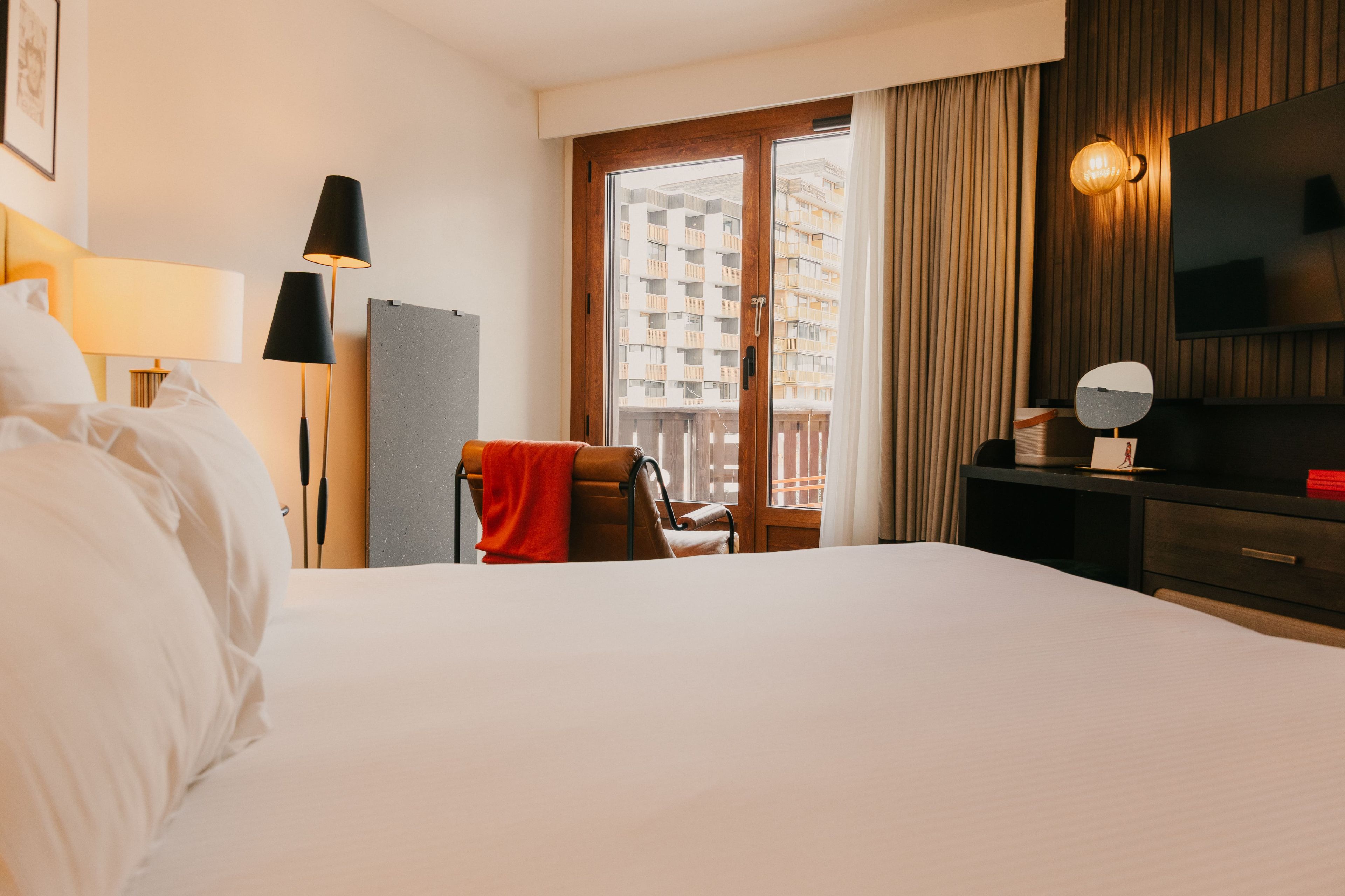 Habitación de hotel con cama, escritorio, silla, televisor y ventana con vistas a edificios.