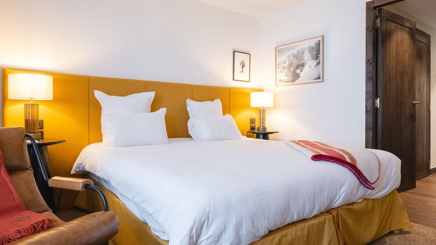A hotel room with a bed, two bedside lamps, a chair, a door, and a framed picture on the wall.