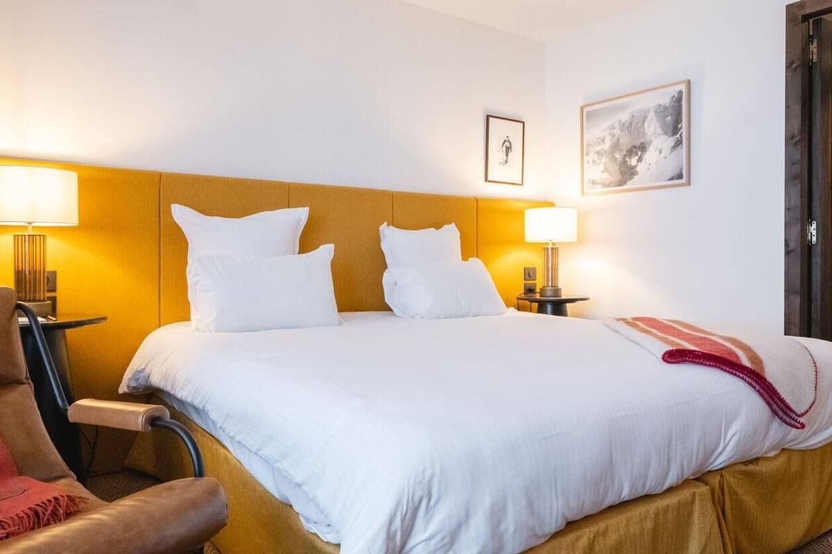 A hotel room with a bed, two bedside lamps, a chair, a door, and a framed picture on the wall.