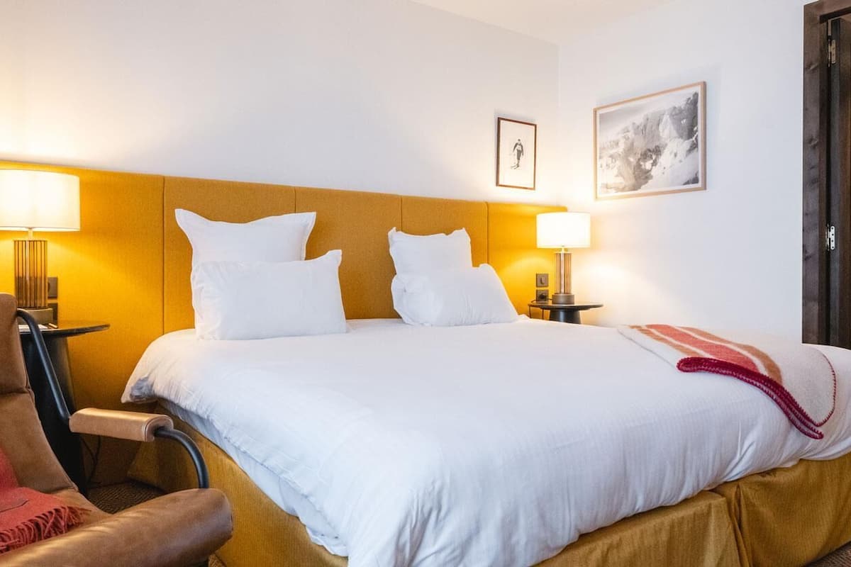 Une chambre d’hôtel avec un lit, deux lampes de chevet, une chaise, une porte et un tableau encadré au mur.