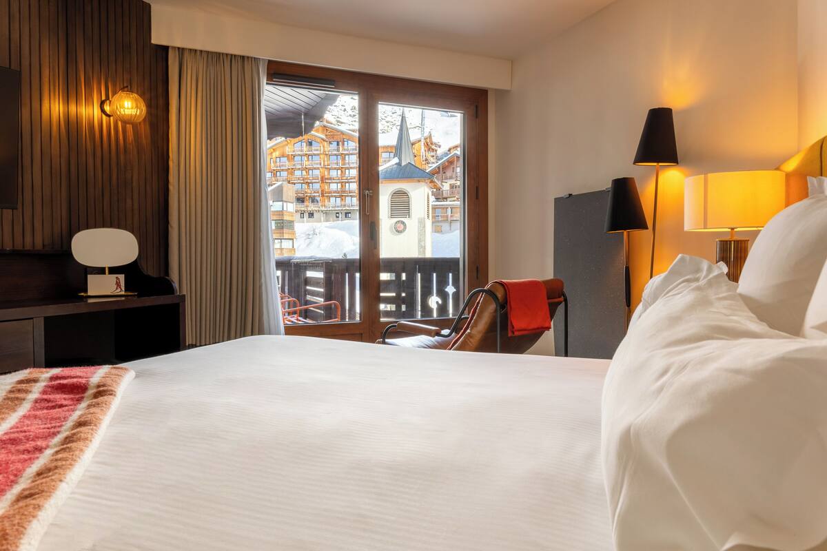 A hotel room with a large bed, a desk, a chair, a lamp, and a view of a building through a window.