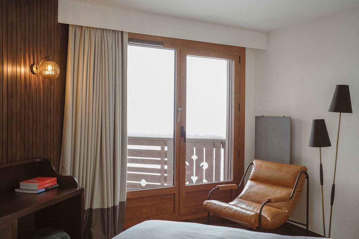 A hotel room with a bed, a desk with books, a leather chair, a balcony with a railing, and a lamp.