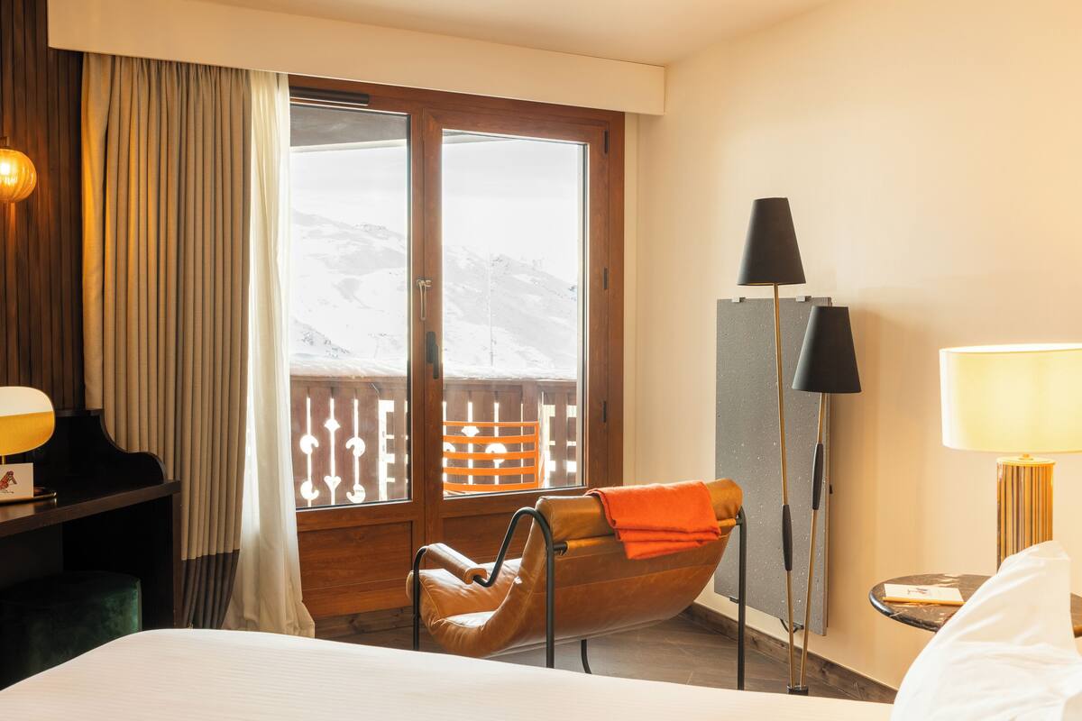 A hotel room with a bed, a chair, a desk, a lamp, and a window with a view of a snowy landscape.