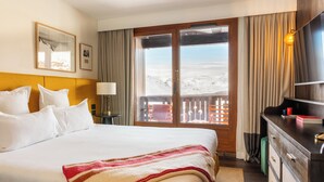 A hotel room with a large bed, a wooden headboard, a desk with a lamp, a dresser, and a view of a snowy landscape through a sliding door. - Le Val Thorens (Les Belleville)