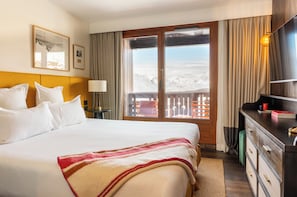A hotel room with a large bed, a wooden headboard, a desk with a lamp, a dresser, and a view of a snowy landscape through a sliding door. - Le Val Thorens (Les Belleville)