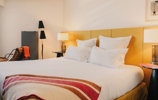 Ein Hotelzimmer mit einem Bett, einem Nachttisch, einer Lampe, einem Sessel und einem an der Wand hängenden Bild.