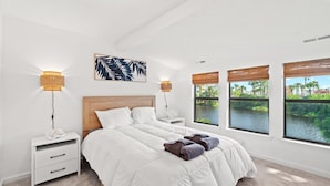 Ein Schlafzimmer mit einem großen Bett, zwei Nachttischen, einer Wandlampe und einem Fenster mit Blick auf ein Gewässer und viel Grün.