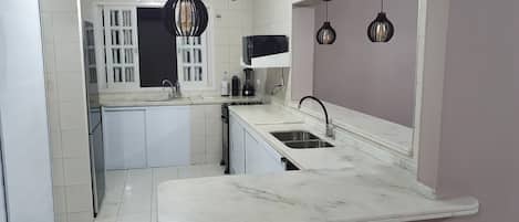 Una cocina compacta con armarios blancos, encimera de mármol, horno empotrado e iluminación colgante.