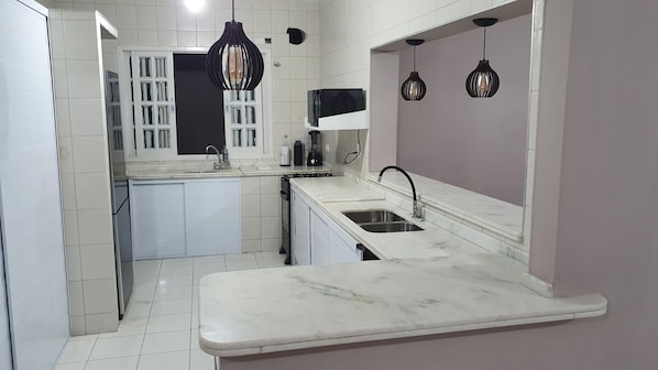 Cozinha compacta com armários brancos, bancada de mármore, forno embutido e iluminação pendente.
