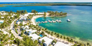 An aerial view of a coastal resort with multiple buildings, a marina with boats, and clear turquoise waters.