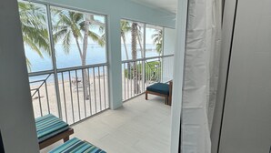 A room with a view of the beach and palm trees, a sliding door, a bed with striped bedding, and a wooden bench. - Kaibo Kove (Rum Point)
