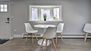 Un comedor con una mesa redonda y cuatro sillas blancas, un ventanal con una planta en maceta, y una puerta con pomo.
