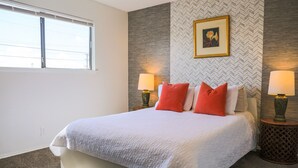 Une chambre à coucher comprenant un lit, deux lampes de chevet, une fenêtre, un tableau accroché au mur et un papier peint à motifs.