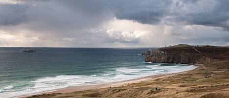 A coastal landscape with a beach, grassy terrain, and rocky cliffs under a cloudy sky.