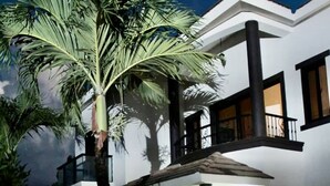 A two-story house with a balcony, a well-lit entrance, and a garden with a palm tree.