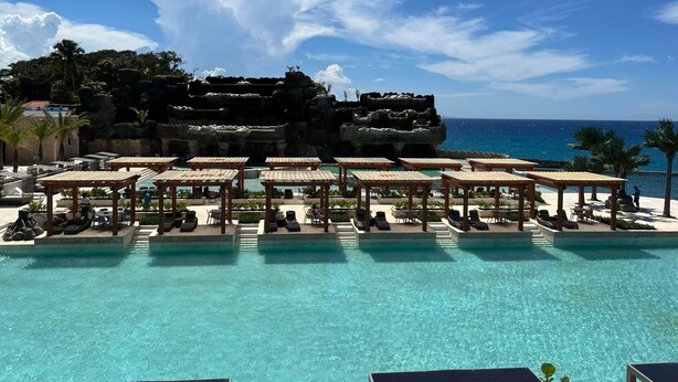 A resort pool area with multiple lounge chairs and umbrellas, a clear view of the ocean, and a rocky cliff in the background.