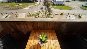 A balcony with a wooden table and chairs, a small garden area with plants and a tree, and a view of a street with cars and buildings. - Modern 3-bedroom townhouse in central Cape Woolamai (Cape Woolamai)