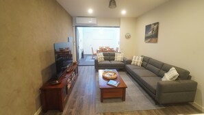 A living room with a sofa, coffee table, and television. There is a dining area with chairs and a table in the background. - Modern 3-bedroom townhouse in central Cape Woolamai (Cape Woolamai)
