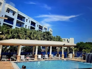 A swimming pool with lounge chairs and a covered area, surrounded by palm trees and a multi-story building in the background. - Updated rates:Oceanwalk bldg 2: 2 bed 2 bath w Ocean view from balcony. Pet fee (New Smyrna Beach)