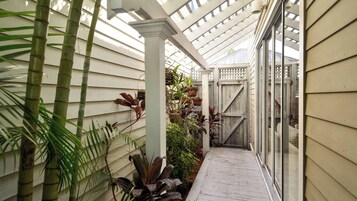 Un corredor exterior cubierto con suelo de madera, paredes de revestimiento blanco y diversas plantas en macetas.
