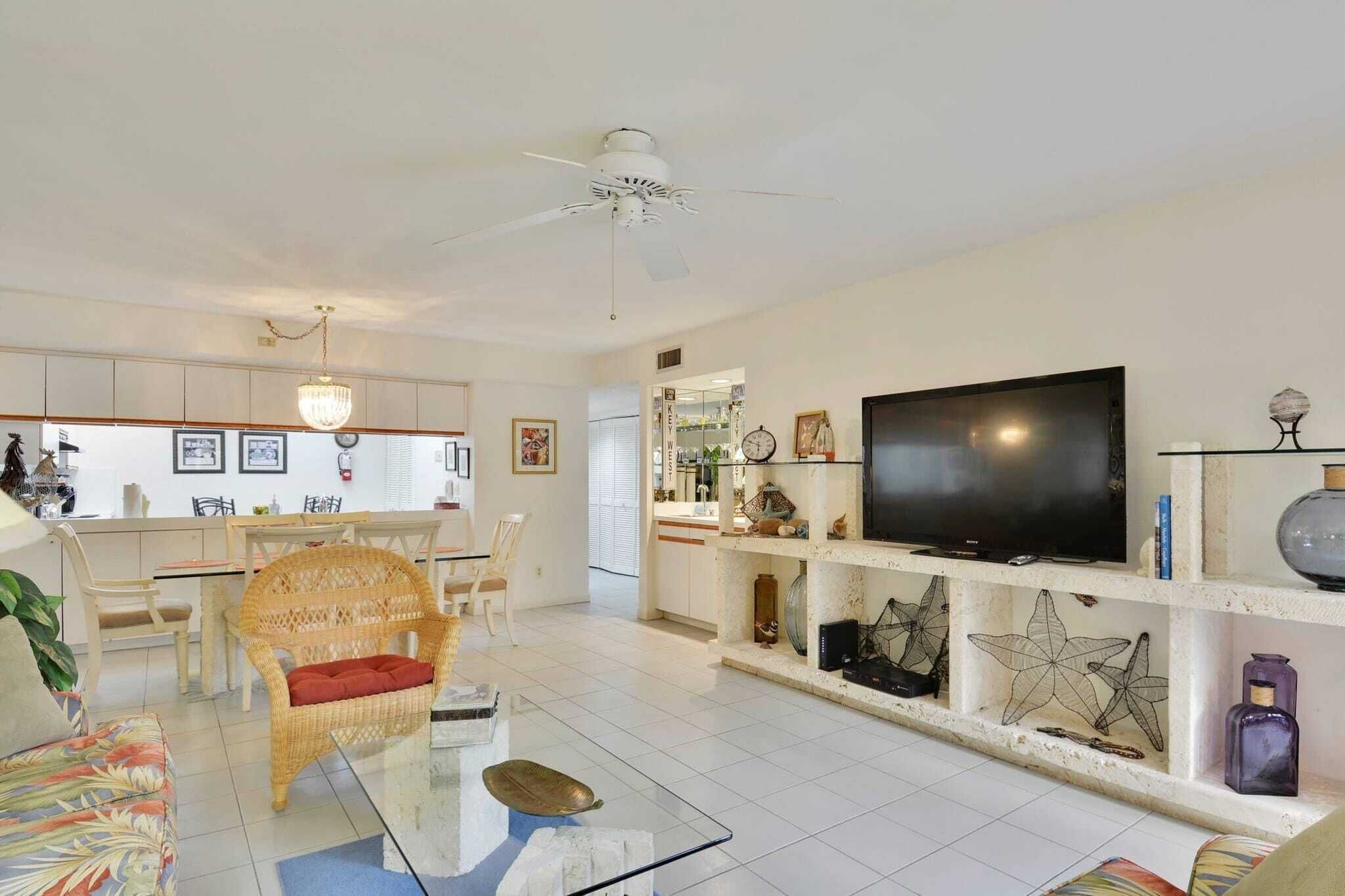 Sala de estar con televisor, muebles de mimbre, una mesa de centro de vidrio y objetos decorativos en estantes.