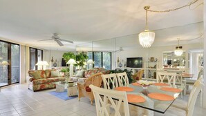 A spacious living room with a glass dining table, white chairs, a sofa, and a ceiling fan. - Atlantic Paradise by Avantstay Great Location w/ Balcony, Outdoor Dining & Shared Pool & Hot Tub! (Key West)