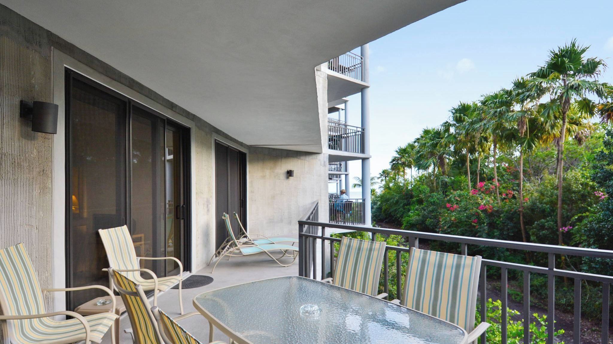 A balcony with a glass table and striped chairs, overlooking a lush garden with palm trees and flowering plants.