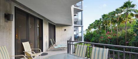 A balcony with a glass table and striped chairs, overlooking a lush garden with palm trees and flowering plants.