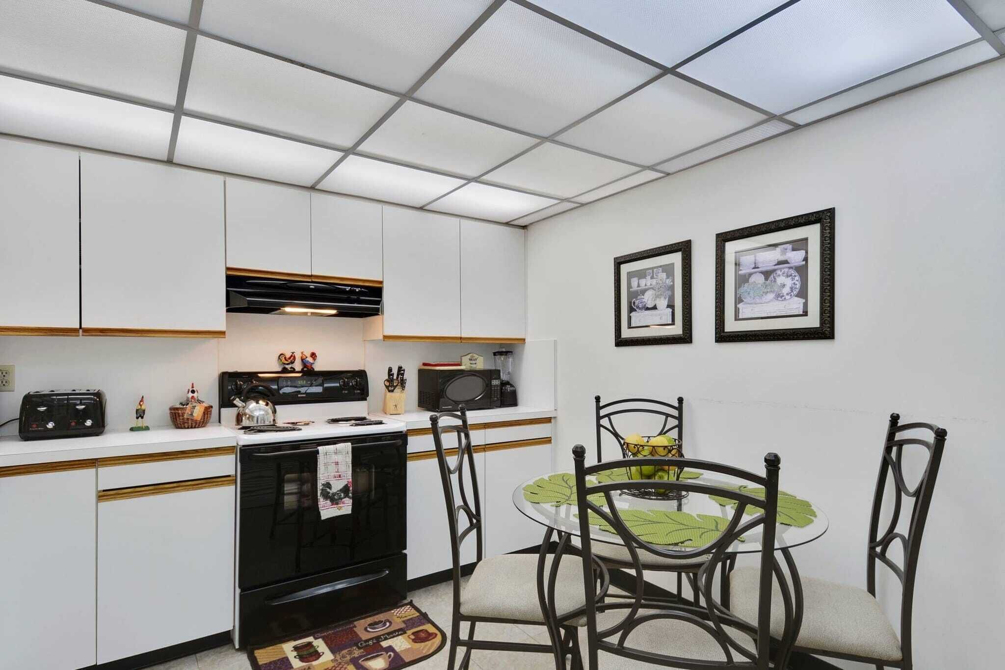 A compact kitchen with white cabinets, a black stove, and a dining table with chairs. There are framed pictures on the wall and a microwave on the counter.