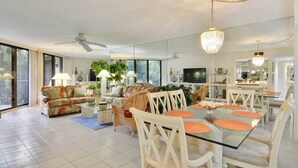 A spacious living area with a glass dining table, white chairs, a sofa, and a ceiling fan. - Atlantic Paradise by Avantstay Great Location w/ Balcony, Outdoor Dining & Shared Pool & Hot Tub! (Key West)