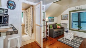 A compact living space with a kitchenette, a grey sofa, a window with blinds, and a decorative wall hanging. - Windchime by Avantstay Walk to Duval Street Month Long Stays Only (Key West)