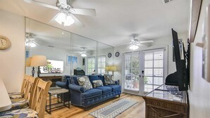 A living room with a blue sofa, wooden chairs, a glass coffee table, and a television. - Always Your Paradise by Avantstay Shared Pool Near Zachary Beach! Month-long Stays Only (Key West)