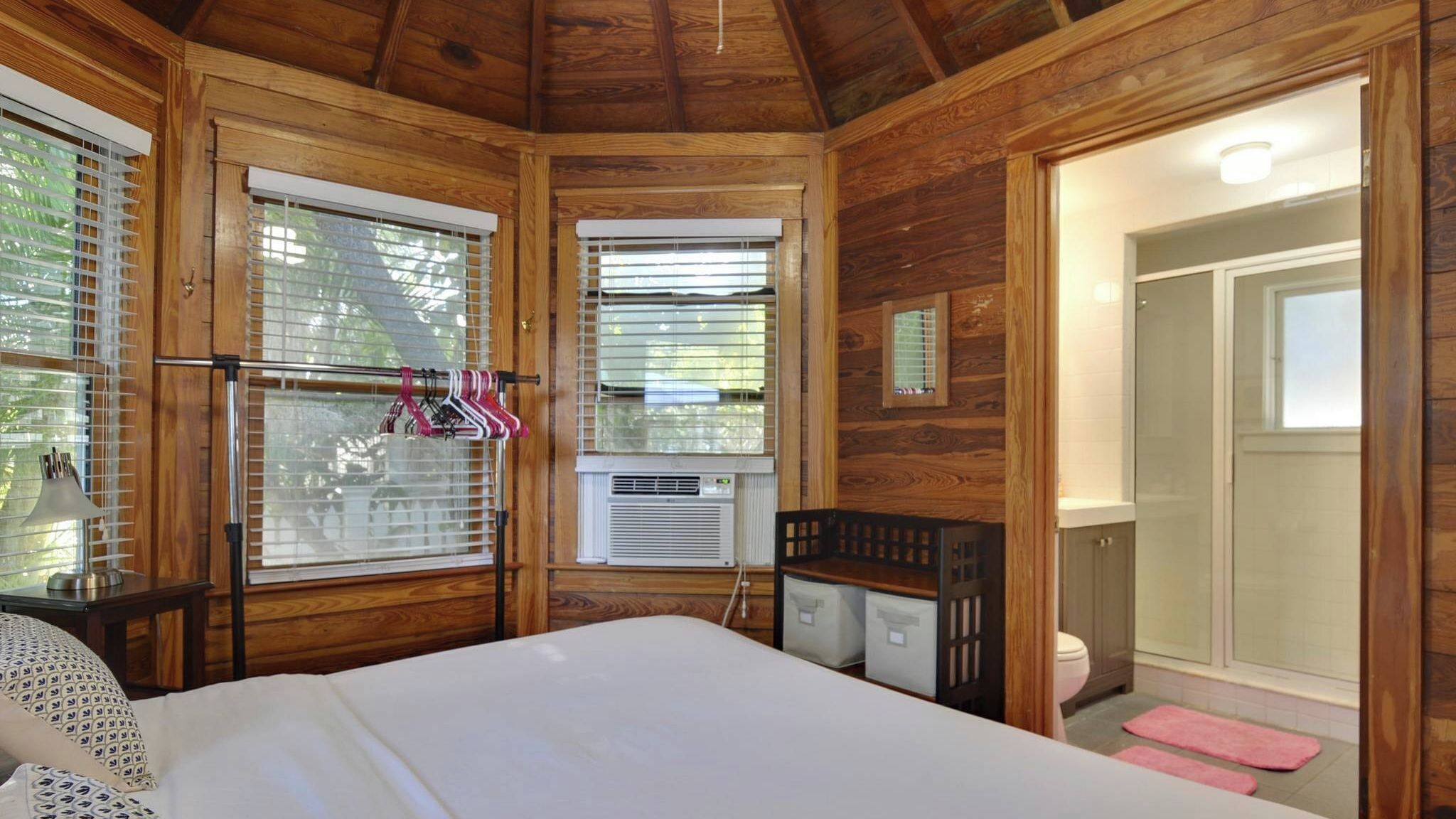 Une chambre aménagée dans une cabane en bois, avec un lit, une petite table et une chaise. Elle est dotée de deux fenêtres équipées de stores, d’un appareil de climatisation et d’une porte donnant sur une salle de bain.