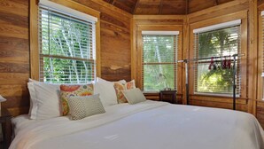 Quarto de cabana de madeira com cama, almofadas, mesa de cabeceira e janela com vista para vegetação.
