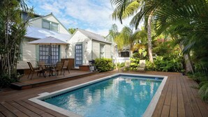 Um quintal com piscina, deck de madeira, móveis de pátio e plantas tropicais.