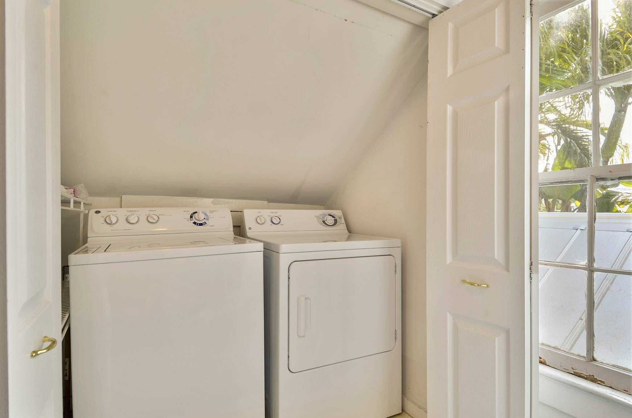 A laundry room with two white washers and dryers, a white door, and a window with a view of palm trees.