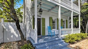 Une maison blanche aux volets verts, un porche avec des chaises bleues et un chemin en briques.