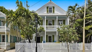 Une maison de style colonial à deux étages, avec une façade blanche, des volets verts et une clôture en bois peinte en blanc.