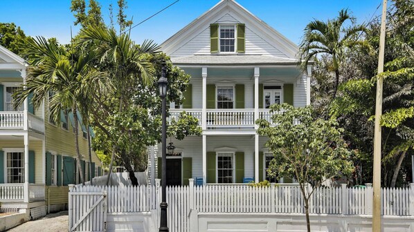 Un bâtiment de style colonial à deux étages, avec une façade blanche, des volets verts et une clôture en bois peinte en blanc.