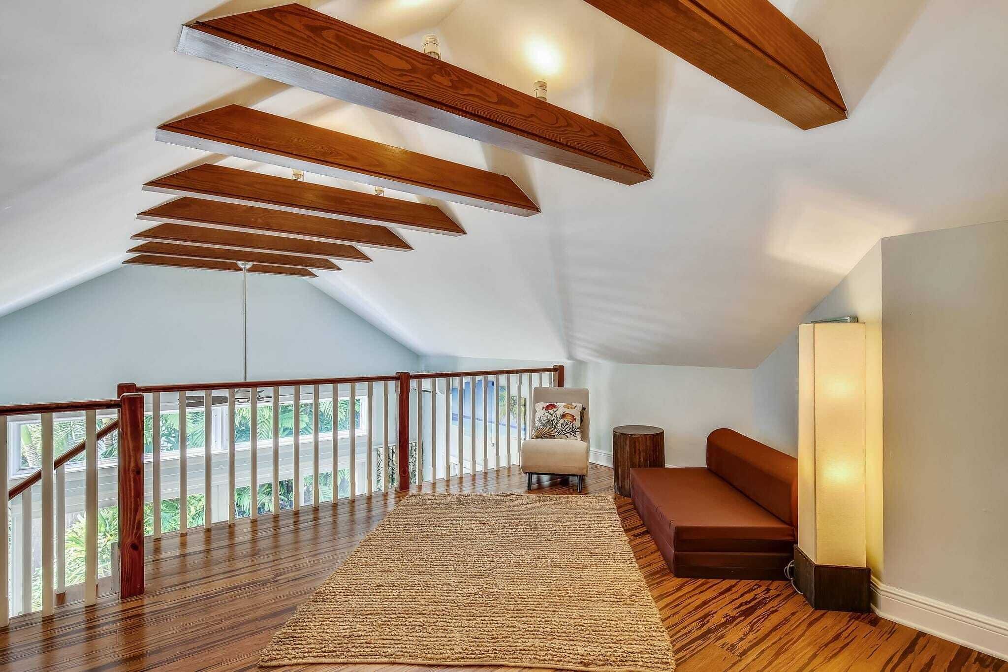 A spacious loft bedroom with a high ceiling, wooden beams, a large window, a cozy seating area, and a view of greenery.
