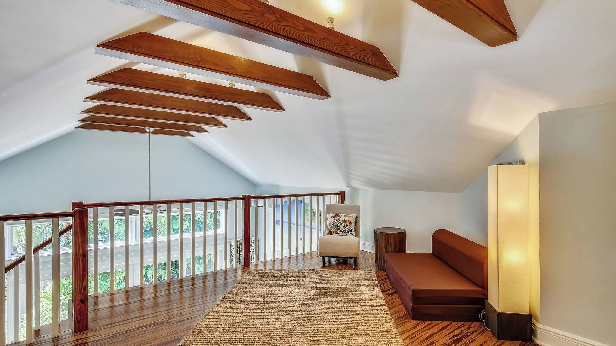 A spacious loft with a high ceiling, wooden beams, a balcony with a view, a cushioned bench, and a floor-to-ceiling window.