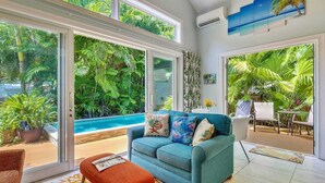 Ampla sala de estar com uma grande porta de vidro deslizante que dá para uma piscina, rodeada por vegetação tropical exuberante.