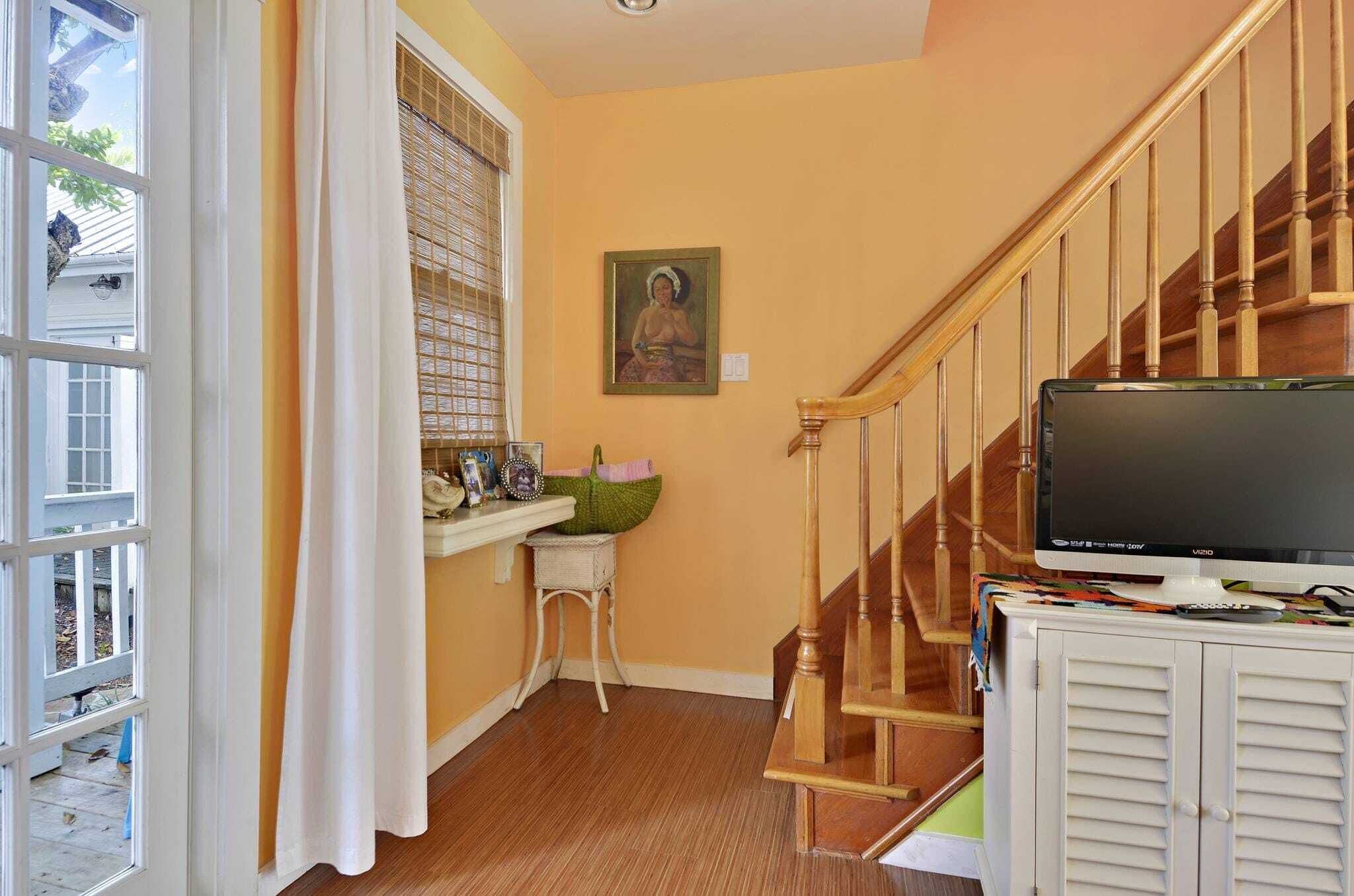 A room with a wooden staircase, a television on a cabinet, a window with blinds, and a small table with decorative items.