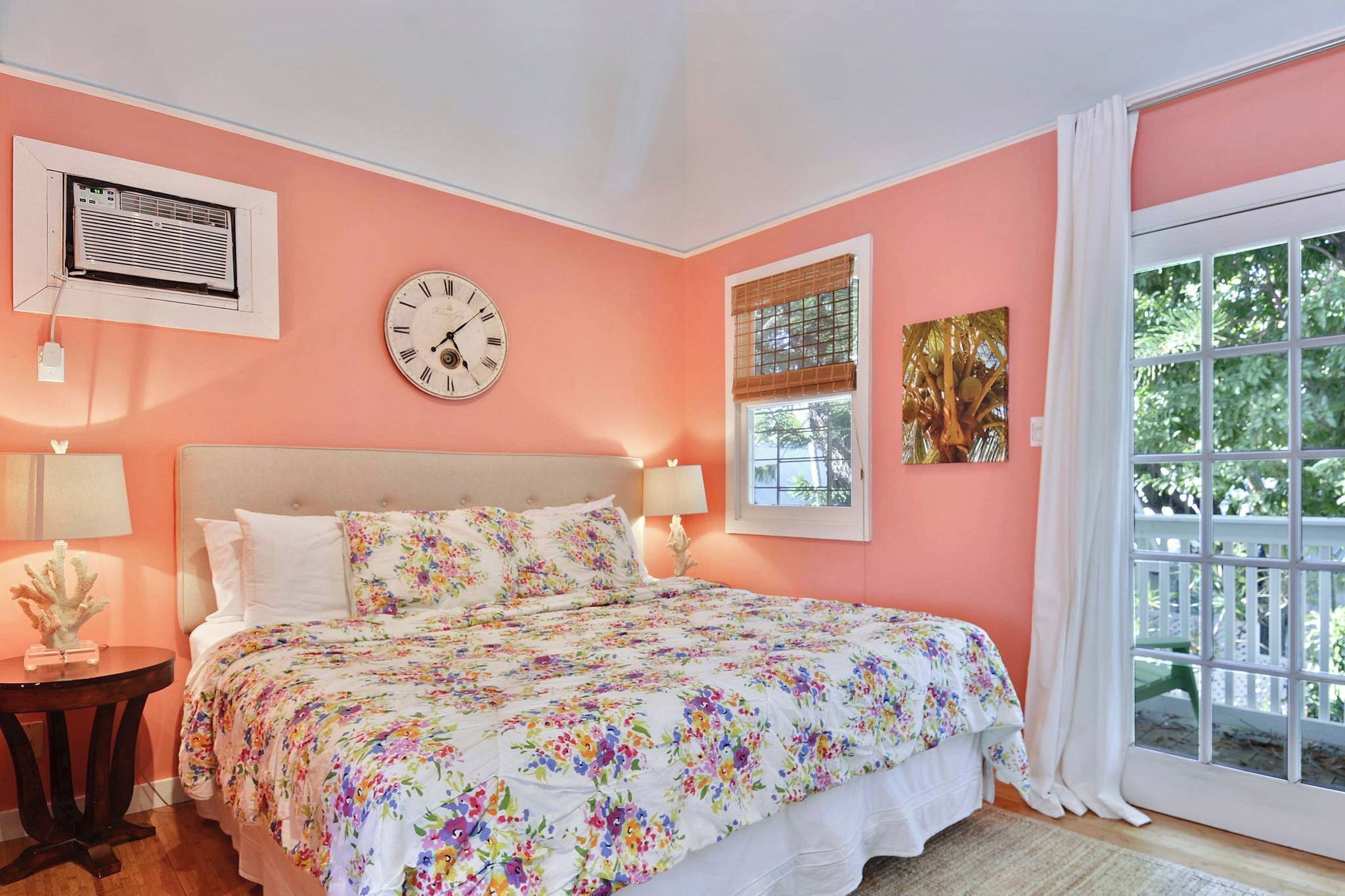 A bedroom with a bed, a clock, an air conditioning unit, a window with a view, and a small table with a lamp.