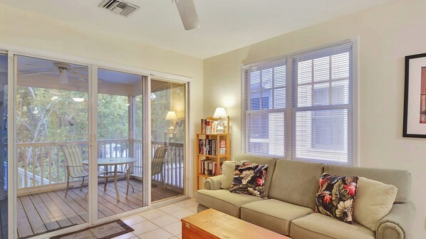A living room with a sofa, a coffee table, a bookshelf, and a sliding glass door leading to an outdoor deck.