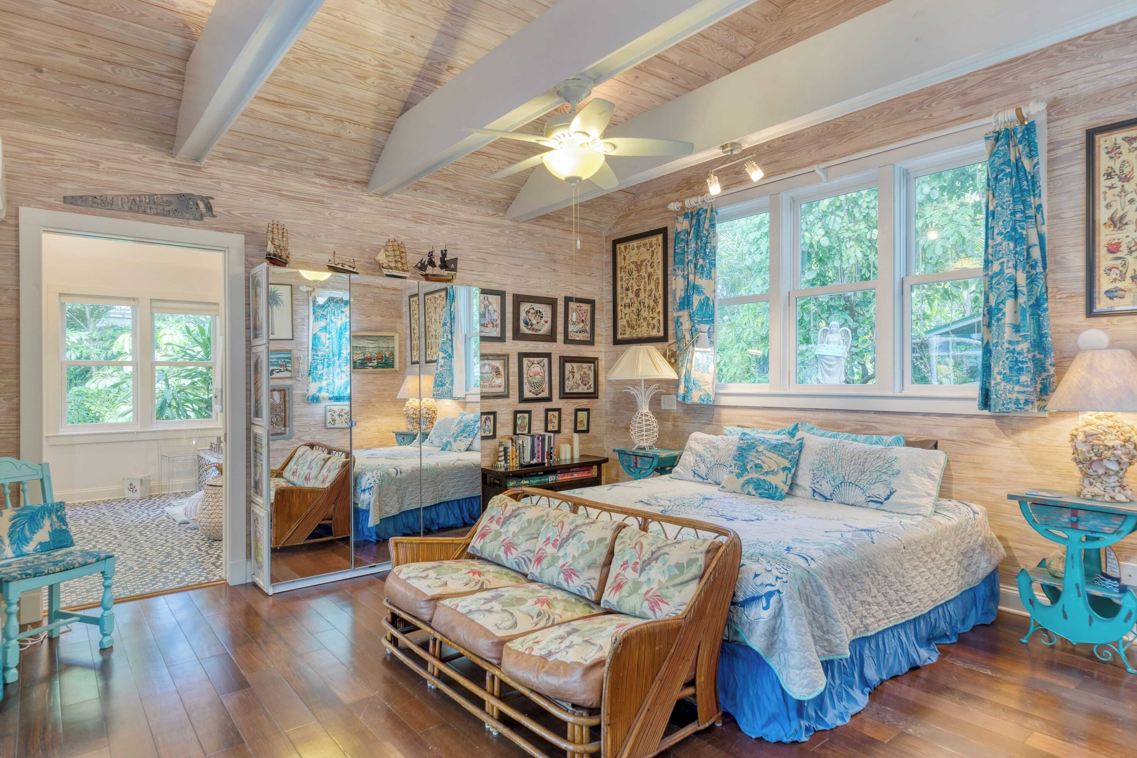 A spacious room with a large bed, a sofa, and a dining table. There are multiple windows providing natural light and a view of the outdoors.