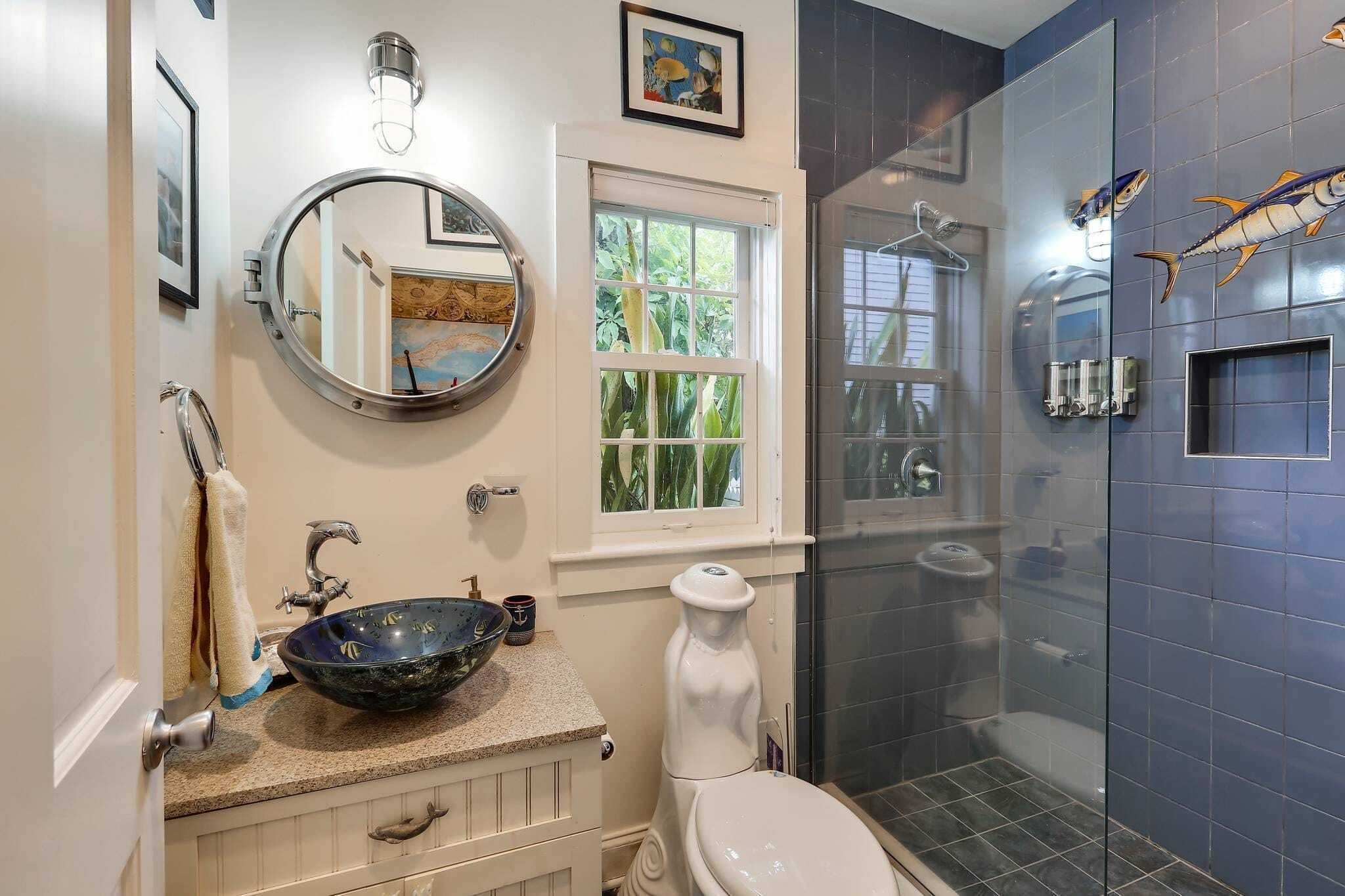 A bathroom with a round mirror, a sink with a decorative bowl, a toilet, a window with a view of greenery, and a shower with a fish mural.