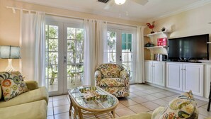 A living room with a sofa, a glass-top coffee table, a patterned armchair, a television, and a bookshelf. - Breezy Bungalow by Avantstay Close 2 Zachary Taylor State Park w/ Shared Pool Month-long Stays (Key West)