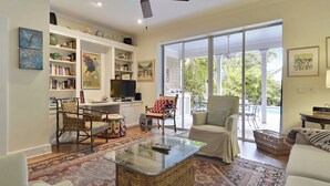 A spacious living room with a large glass door leading to a balcony, a bookshelf filled with books, a television, and a variety of seating options. - Amelia Home by Avantstay In Historic Old Town w/ Pool (Key West)